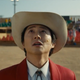 A man (Steven Yeun) wearing a cowboy hat, white shirt with bolo tie, and red jacked looks up at the sky in a scene from Nope.